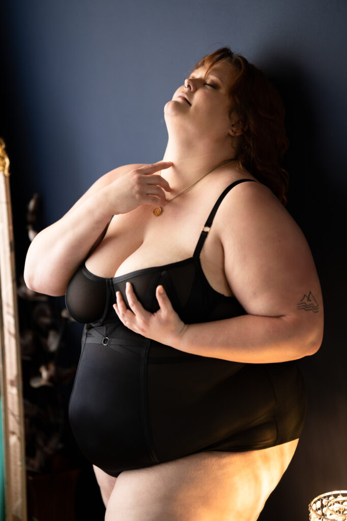 A confident plus-size woman in black lingerie, leaning against a vibrant blue wall.