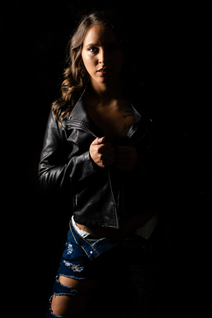 A woman in jeans and a black leather jacket stands in front of a black background.