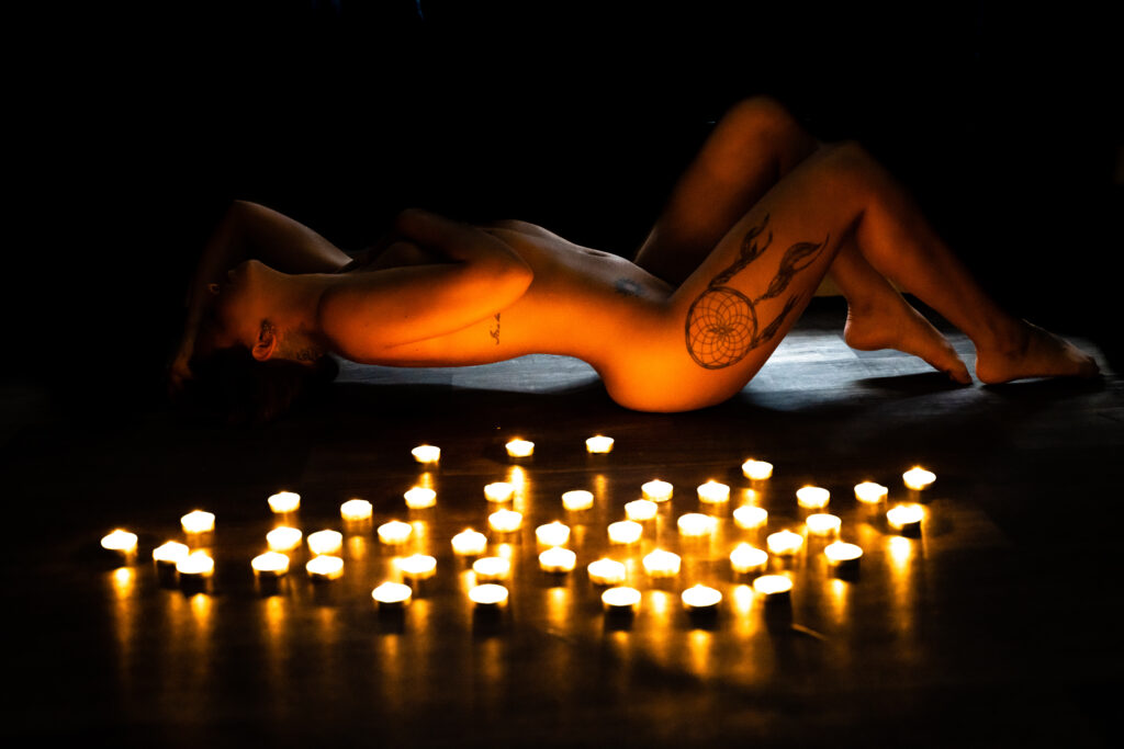 Image of a woman laying down on her back on the floor in front of candles. She is nude with a tattoo of a dreamcatcher on her high. The candles are casting a soft light on her skin, creating a sensual and intimate atmosphere.